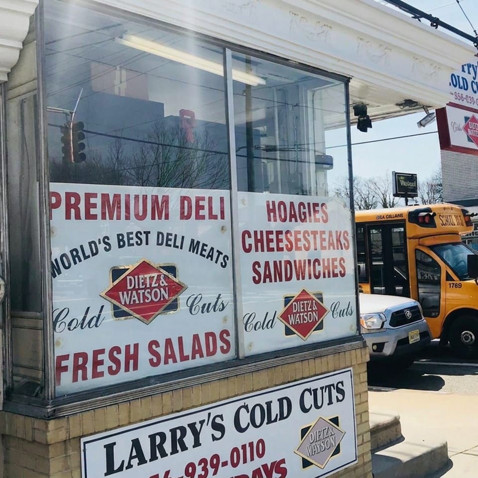Larry’s Cold Cuts deli storefront displays signs for premium meats, hoagies, cheesesteaks, sandwiches, and fresh salads beside a yellow bus.