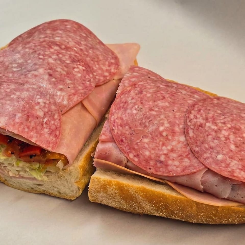 Two open-faced deli sandwiches topped with overlapping salami slices, layered meats, lettuce, tomatoes, and cheese on white bread.