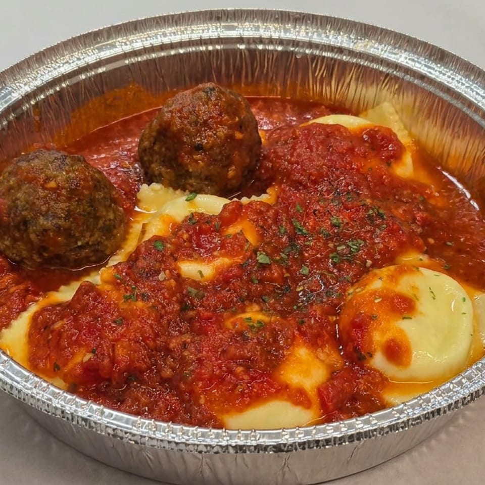 Aluminum tray of meatballs and melted cheese-covered pasta topped with chunky tomato sauce and herbs.