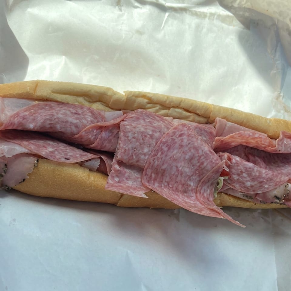 Overstuffed Italian sub with salami, ham, and cheese on a long roll, wrapped in paper beside a bottled drink.
