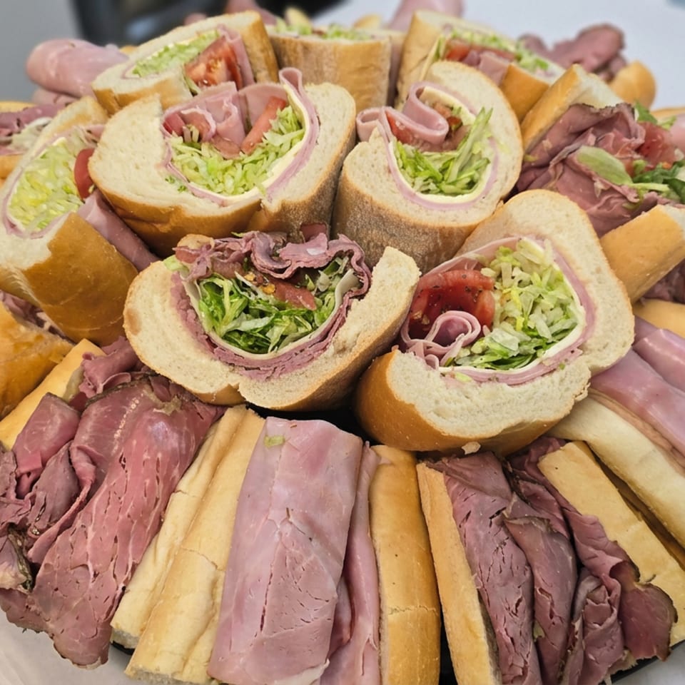 Catering platter featuring sliced deli meat sandwiches with lettuce and tomato, surrounded by assorted meats and bread rolls.