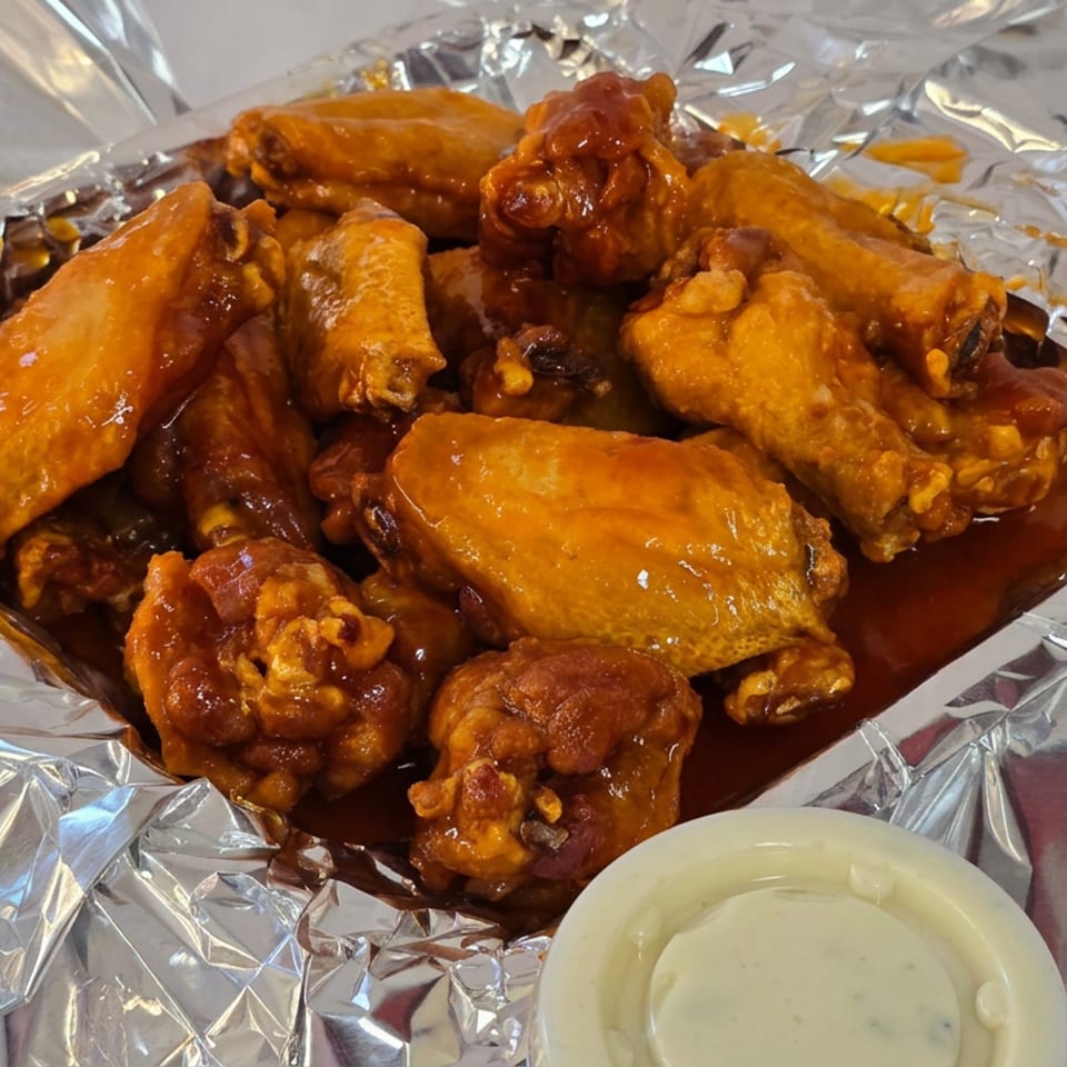 Pile of saucy golden chicken wings served on foil with a cup of creamy dipping sauce.